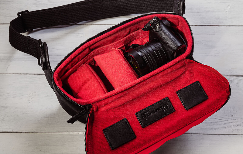 Open red camera bag with black shoulder strap displaying internal padding, dividers, and a compact black camera with lens inside.
