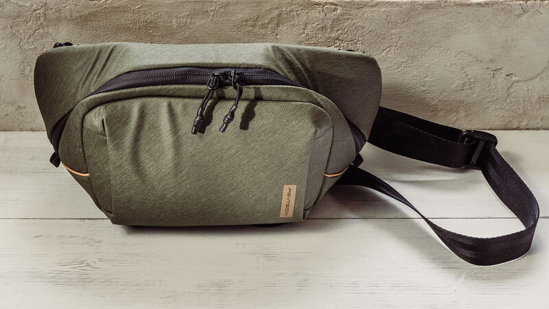 Sage green camera sling bag with black shoulder strap on white wood surface.