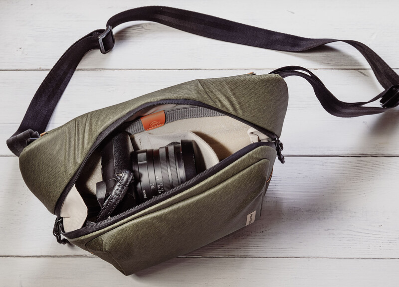 Olive canvas camera bag with black shoulder strap lying open on white wooden surface, revealing a mirrorless camera with attached lens inside.