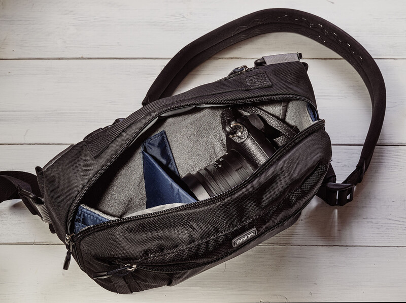 Open black camera bag displaying interior compartments with camera gear, lens, and accessories on white wooden surface.