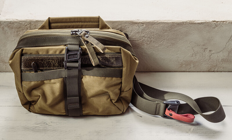 Tan canvas camera bag with brown accents and shoulder strap on white wooden surface.