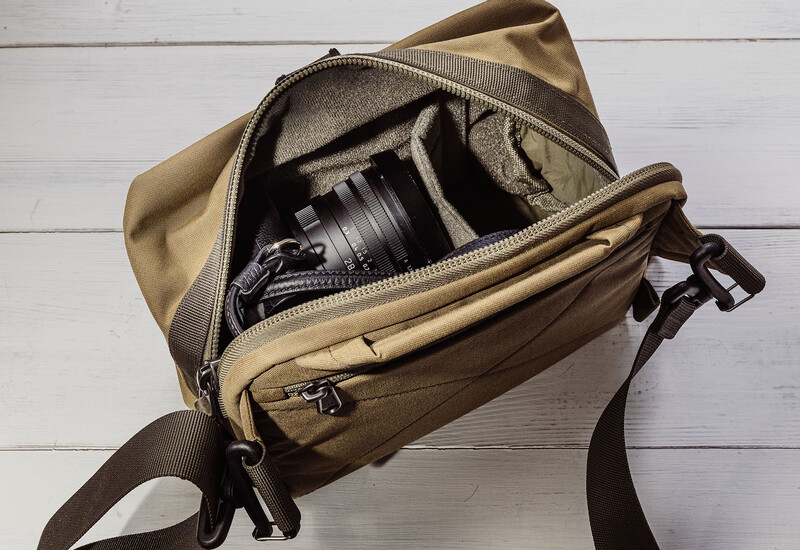 Tan canvas camera bag opened to reveal a mirrorless camera with attached lens and foam compartments on white wooden surface.
