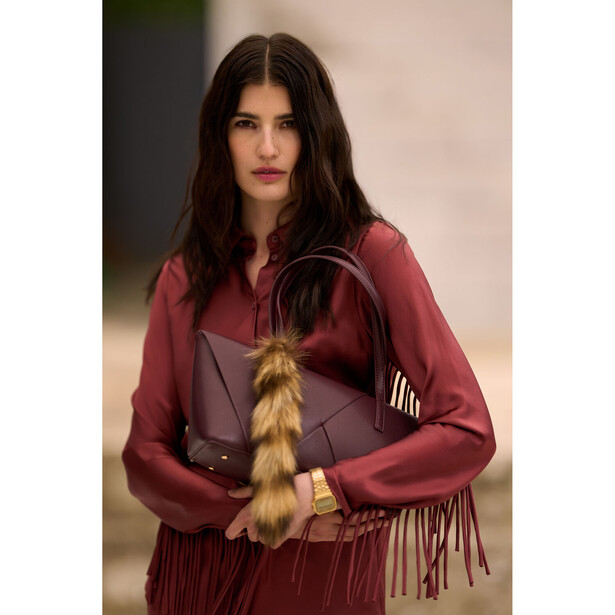 Woman in burgundy shirt holding brown leather handbag with fringe detail against neutral background.