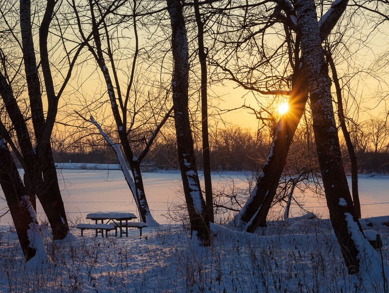 Winter photo of the sun rising over frozen lagoons, with a snow-covered picnic table