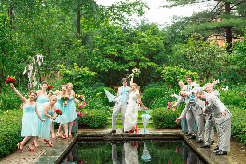 Bride and groom posed on a wooden bridge over a pond with wedding party members lined up on both sides.