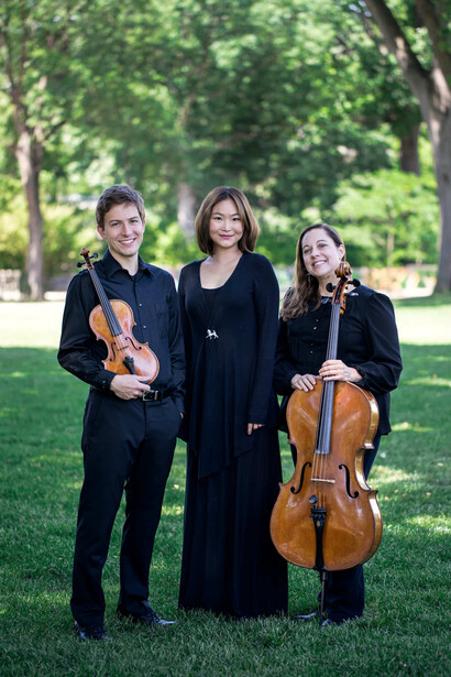 Three musicians in black formal attire holding violin and cello instruments outdoors on grass.