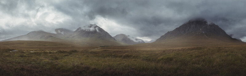 Three snow-capped mountains rise from a barren moorland beneath an overcast sky.