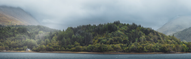 Densely forested mountainous coastline with coniferous trees under overcast skies and a small white structure visible along the water's edge.