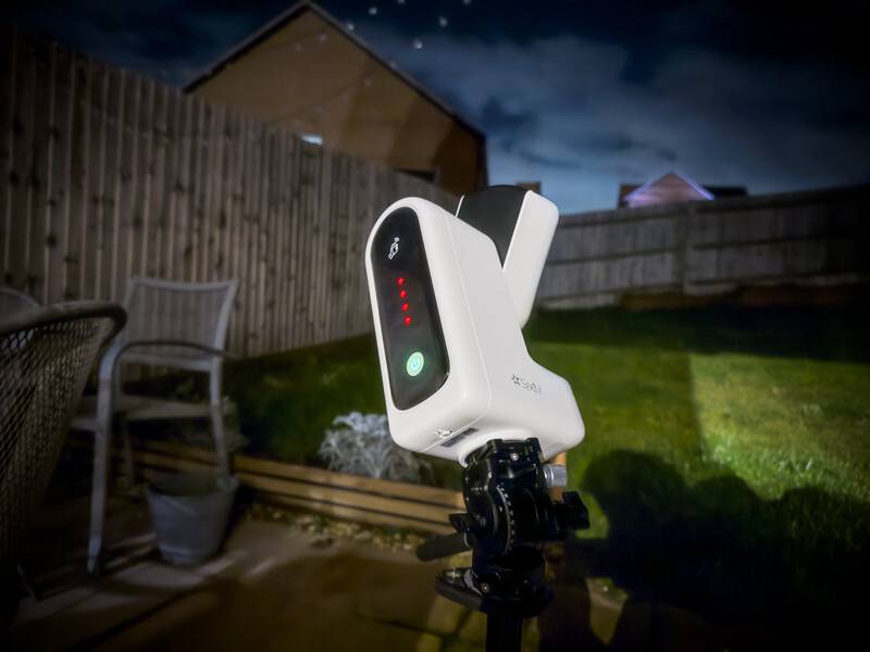 White and black camera flash or lighting device mounted on tripod in backyard at dusk.