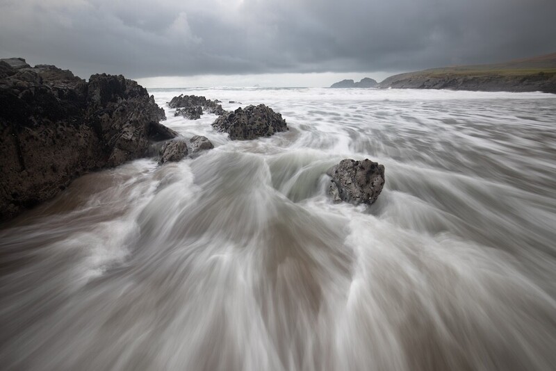 Silky seawater flows around dark rocky outcrops on a windswept coastal beach.