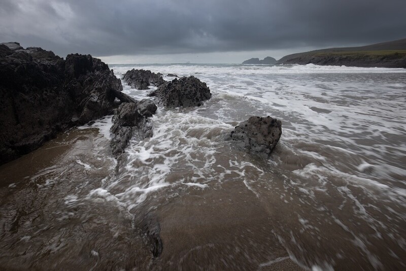 Turbulent waves crash against dark rocky outcrops on a moody coastal beach.