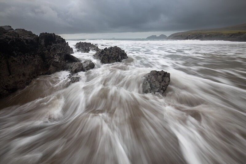 Silky seawater swirls around dark rocks on a beach beneath an overcast sky.