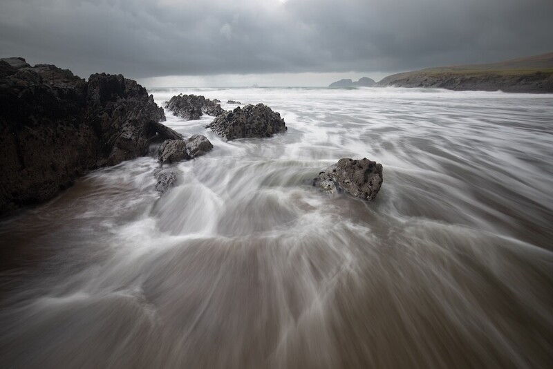 Seascape with silky water flowing around dark rocks under overcast sky.