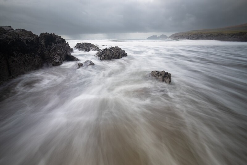 Long exposure photograph of waves flowing around dark rocky outcroppings on a misty beach.