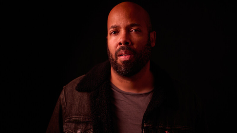 Man with beard in dark denim shirt against black background with warm red lighting.