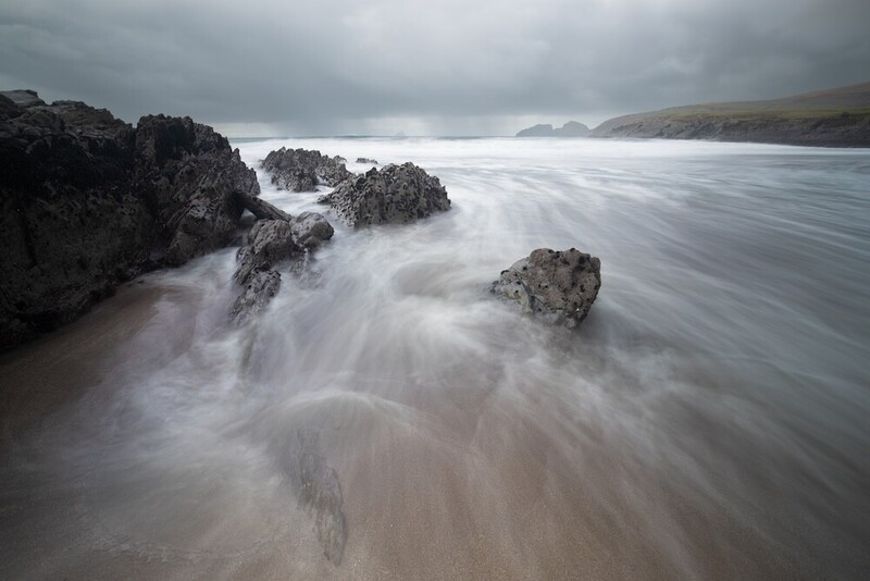 Seascape with silky water flowing around dark rocky outcroppings on a moody beach.