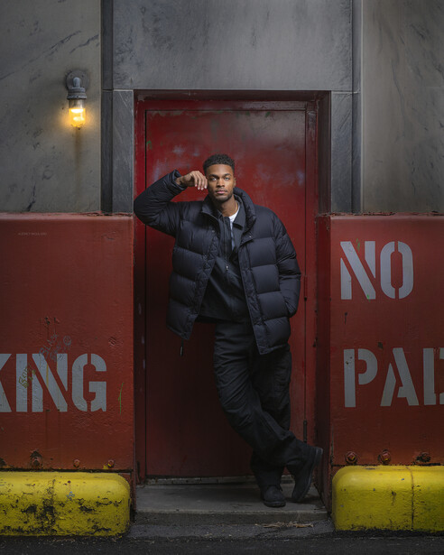 Man in dark puffy jacket leaning against red door framed by yellow bollards and industrial gray walls.