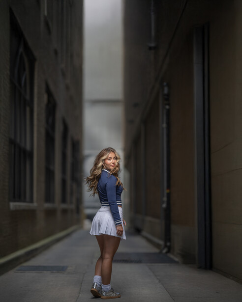 Young girl in blue and white dress standing alone in a narrow urban alleyway between tall dark buildings.