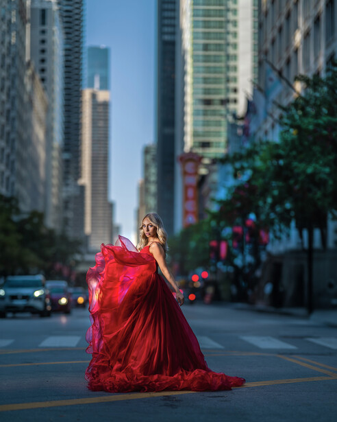 Woman in flowing red gown posing on urban street lined with tall buildings.