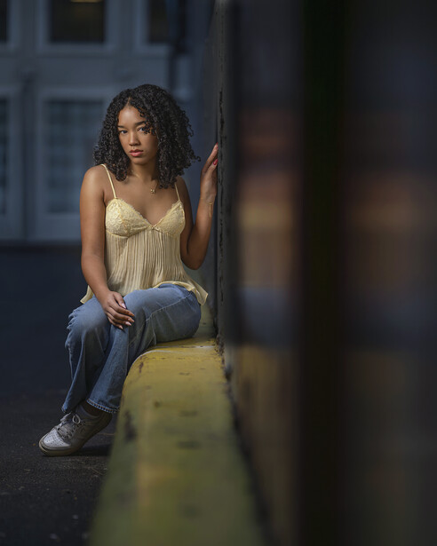 Woman in yellow tank top and jeans sitting on a concrete ledge in an urban alley at dusk.