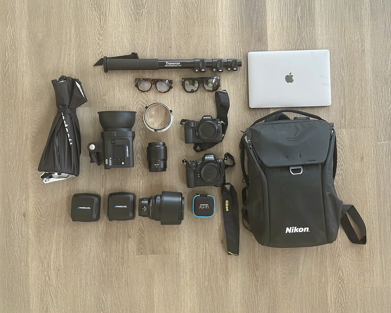 Flat lay of professional photography gear including Nikon camera body, lenses, MacBook, backpack, and accessories on wood floor.
