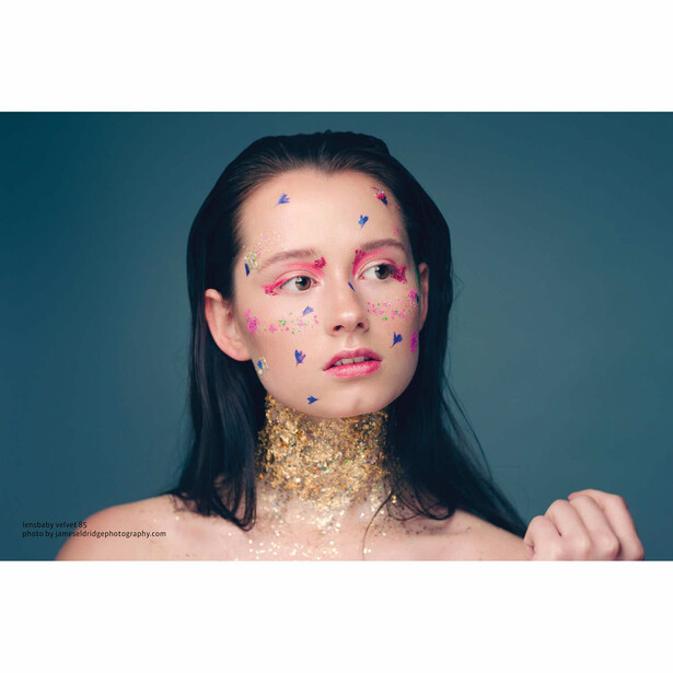 Portrait of a woman with long dark hair against a teal background, decorated with colorful geometric shapes on her face and neck.