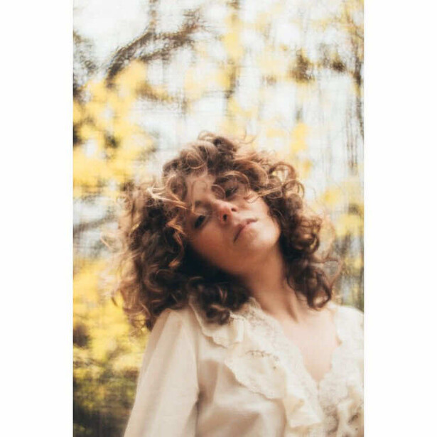Person with curly reddish-brown hair tilted back, wearing a cream-colored shirt, with dappled sunlight and blurred foliage in background.