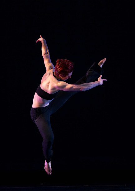 Dancer suspended in mid-air performing a high kick with arms extended against a black background.
