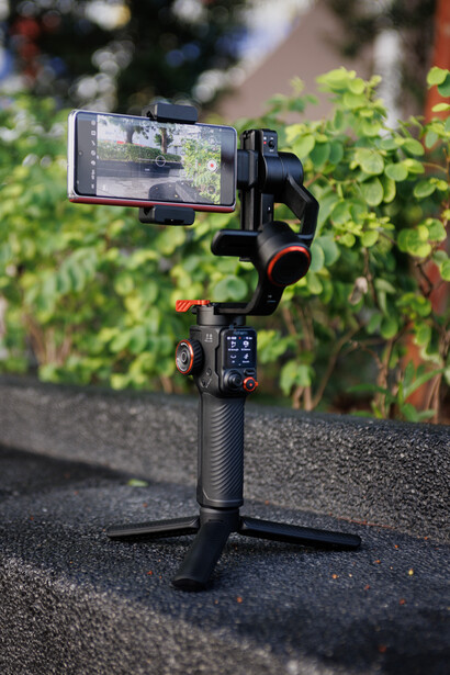 Three-axis gimbal stabilizer mounted on tripod with smartphone displaying live video feed outdoors.
