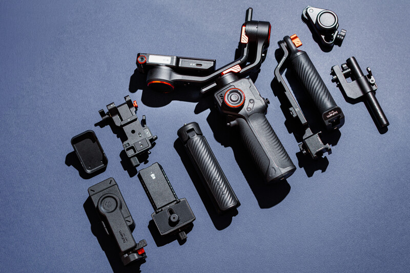 Flat lay of video production equipment including gimbal stabilizer, wireless microphones, batteries, and accessories arranged on blue background.