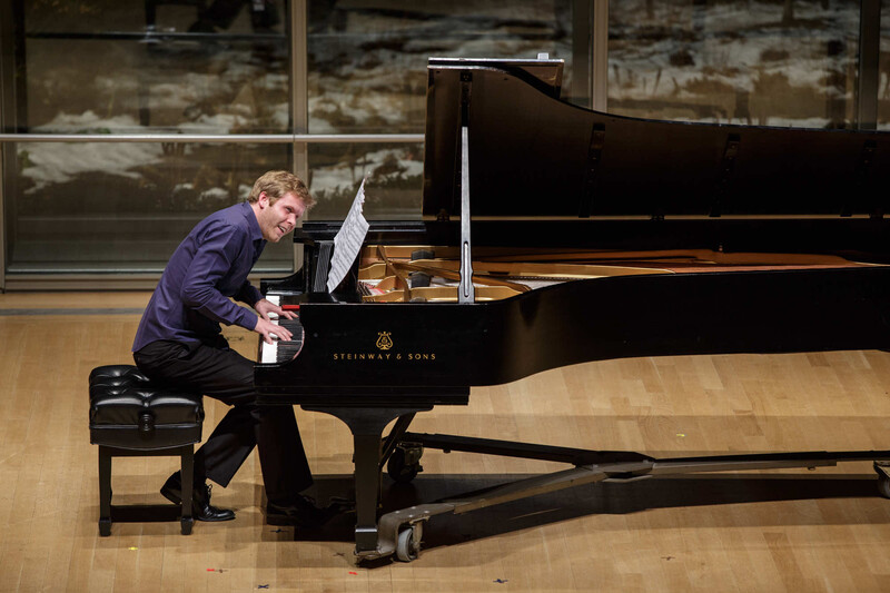 Pianist performing at a grand piano on a wooden stage with large windows behind.