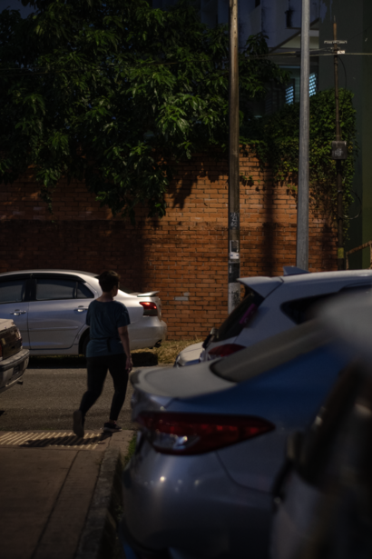 Person walking between parked cars on an urban street at dusk, with brick building and vegetation in background.