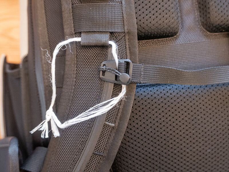 Close-up of a camera backpack's shoulder strap attachment with a carabiner clip and white cord detail.