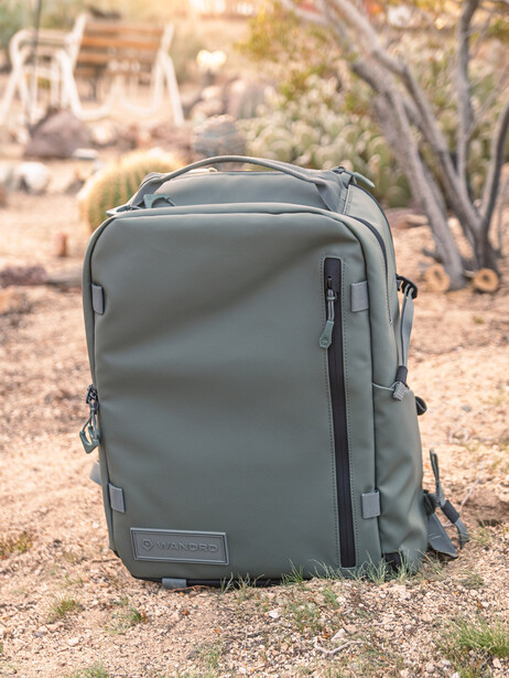 Gray travel backpack with multiple compartments sitting on sandy ground outdoors.