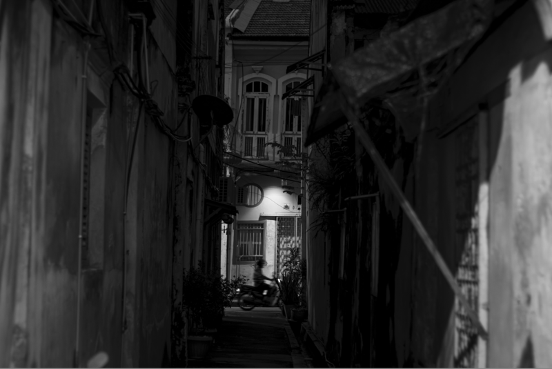 Narrow alleyway at night with a lone figure seated in the distance under a streetlight.