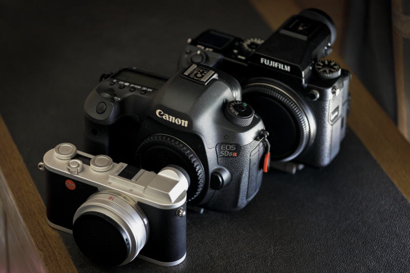 Three cameras arranged on a dark surface: a silver rangefinder, a black Canon EOS DSLR, and a black Fujifilm mirrorless camera.