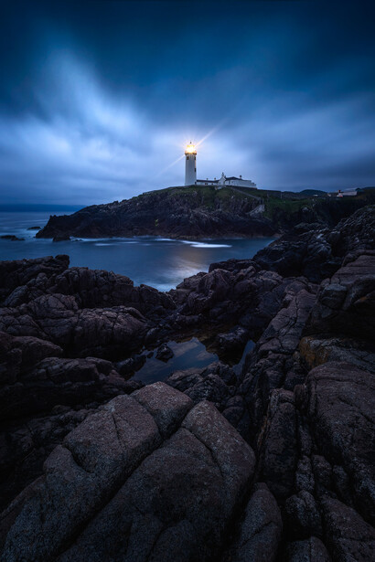 Illuminated lighthouse on a rocky coastal peninsula at twilight with stormy clouds overhead.