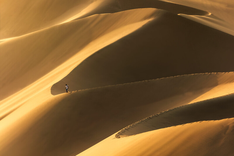 Lone figure walking across undulating sand dunes with dramatic shadows and golden light.