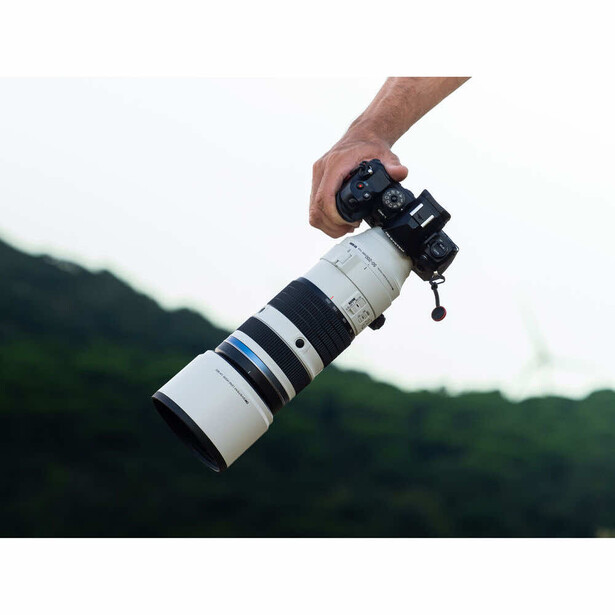 Hand holding a mirrorless camera with a white telephoto lens against a blurred green mountainous background.