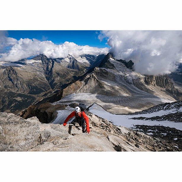 Paraglider in red harness launching from alpine ridge with snow-capped peaks and glacier-fed lake below.