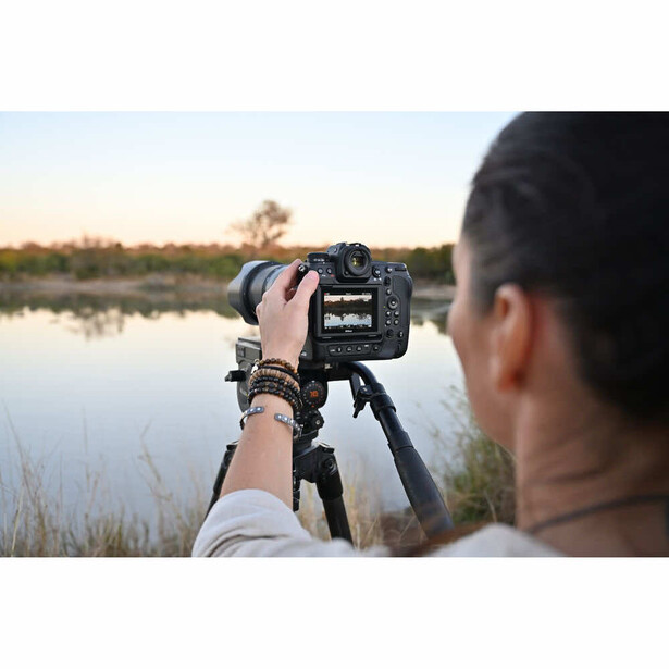 Photographer shooting landscape with mirrorless camera on tripod beside still water at sunrise.
