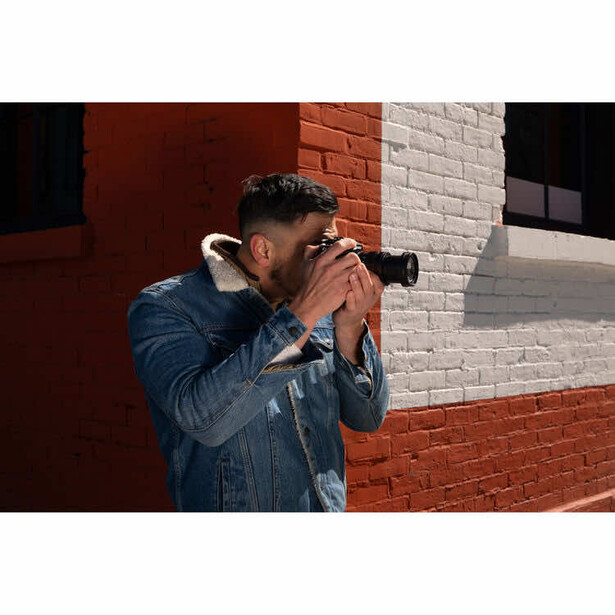 Male photographer holding a telephoto lens camera to his face while standing against a brick wall.