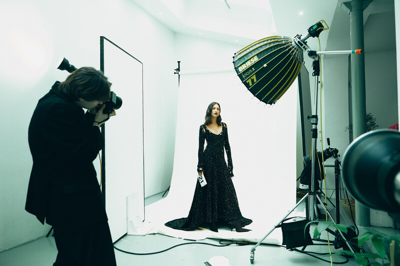 Photographer operating a camera on a tripod while a model in a black dress poses in a studio lit by a large overhead light fixture.