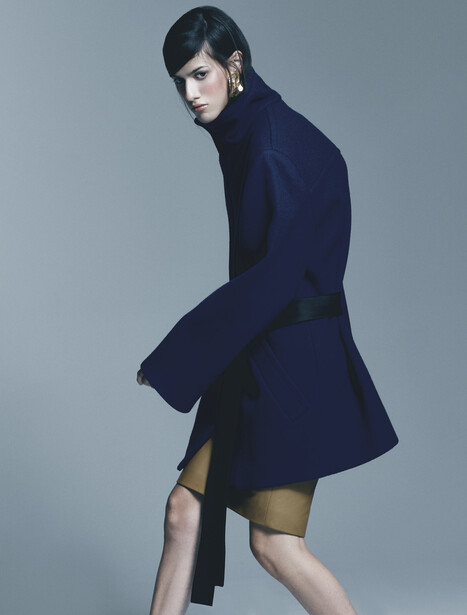 Woman in navy blazer dress posed against neutral gray background, shot from side angle.