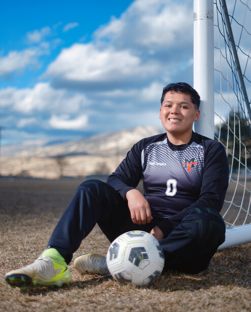 Young soccer player sitting on ground against goal net holding a soccer ball.