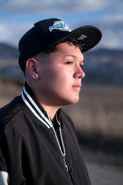 Young person in profile wearing a black cap and dark jacket with lanyard, photographed outdoors with blurred landscape background.