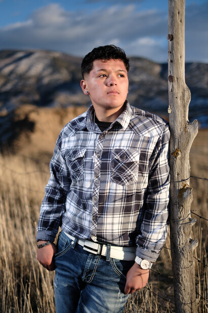Young man in plaid shirt standing against wooden post in dry grassland with mountains in background.