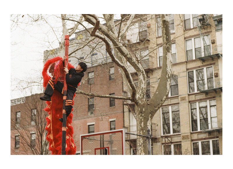 Person climbing a bright red vertical installation on a city street lined with bare trees and brick buildings.