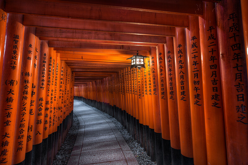 Tunnel of bright orange torii gates with black calligraphic text receding into the distance.
