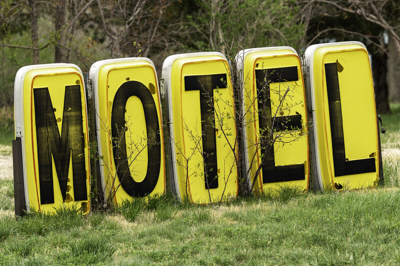 Five yellow motel signs with black lettering arranged in a row on grass with bare trees in background.